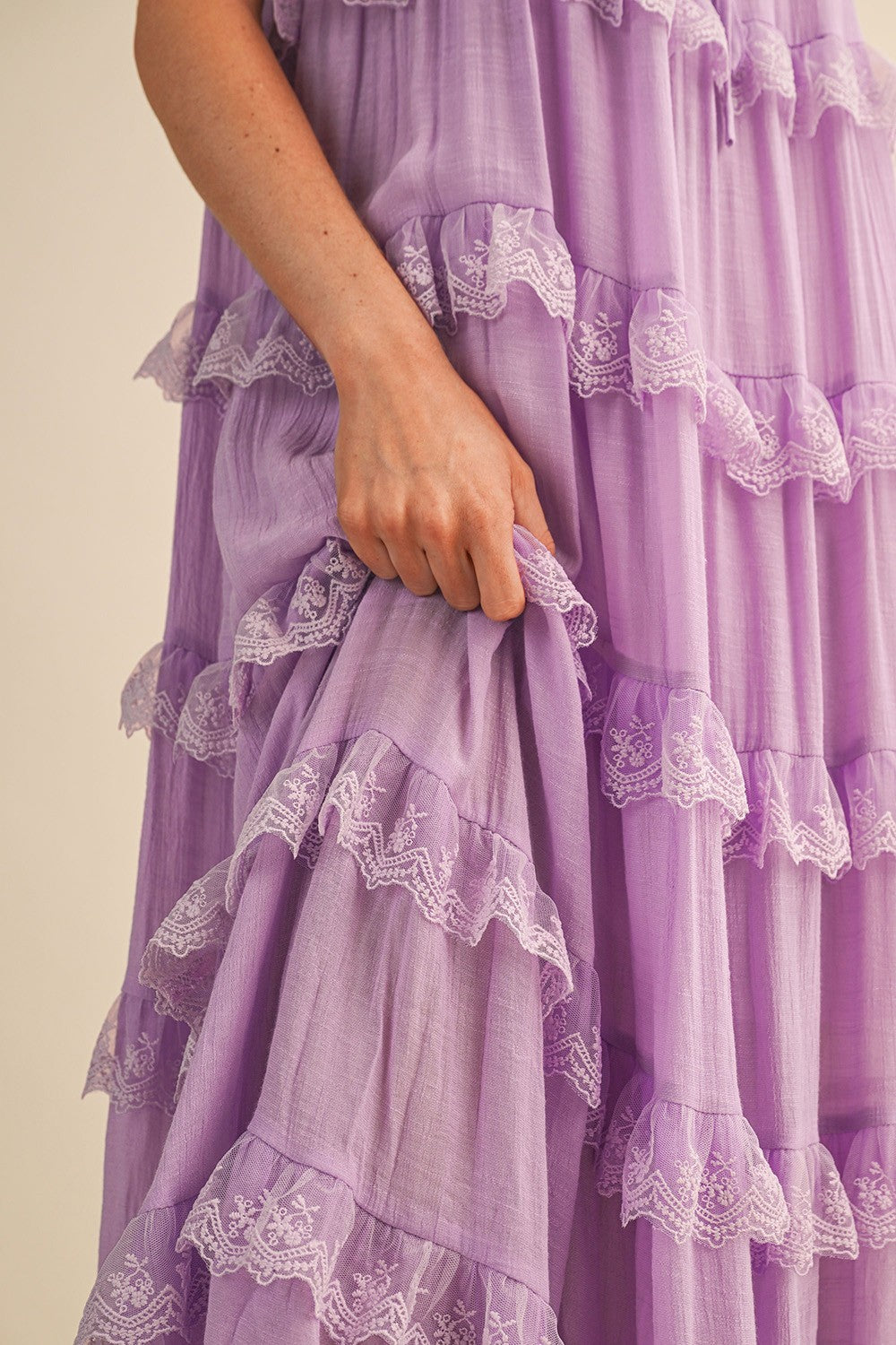 A sleeveless tiered maxi dress in lavender featuring intricate scalloped lace trim, a scooped neckline with an adjustable front tie, and flutter shoulder straps.