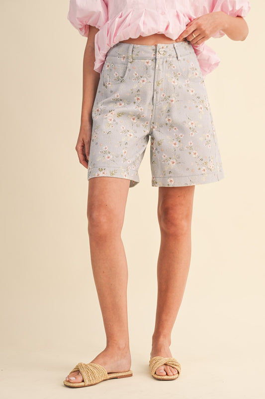 A pair of high-waisted women's light denim shorts in a light wash with a delicate pink and green floral print, featuring a modest mid-length hem and classic front pockets.