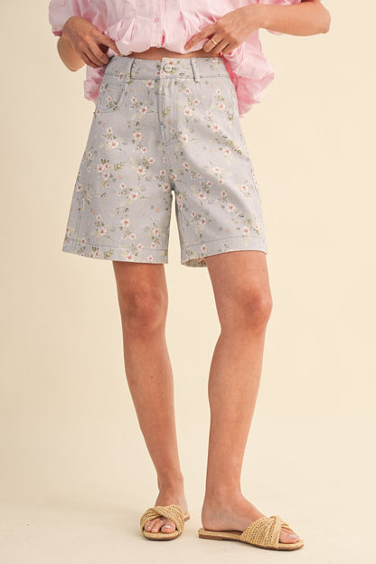 A pair of high-waisted women's light denim shorts in a light wash with a delicate pink and green floral print, featuring a modest mid-length hem and classic front pockets.