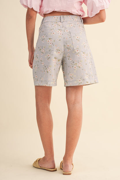 A pair of high-waisted women's light denim shorts in a light wash with a delicate pink and green floral print, featuring a modest mid-length hem and classic front pockets.