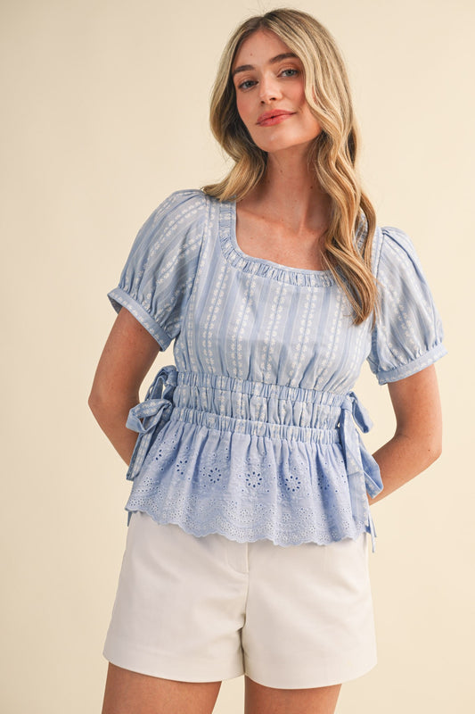 Shirred scoop neckline and tiered elastic waistband blouse. With short puff sleeves, buttoned cuffs, delicate bow ties at the sides, and a striped floral eyelet peplum hem in cornflower blue. 