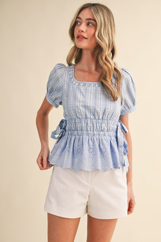Shirred scoop neckline and tiered elastic waistband blouse. With short puff sleeves, buttoned cuffs, delicate bow ties at the sides, and a striped floral eyelet peplum hem in cornflower blue. 