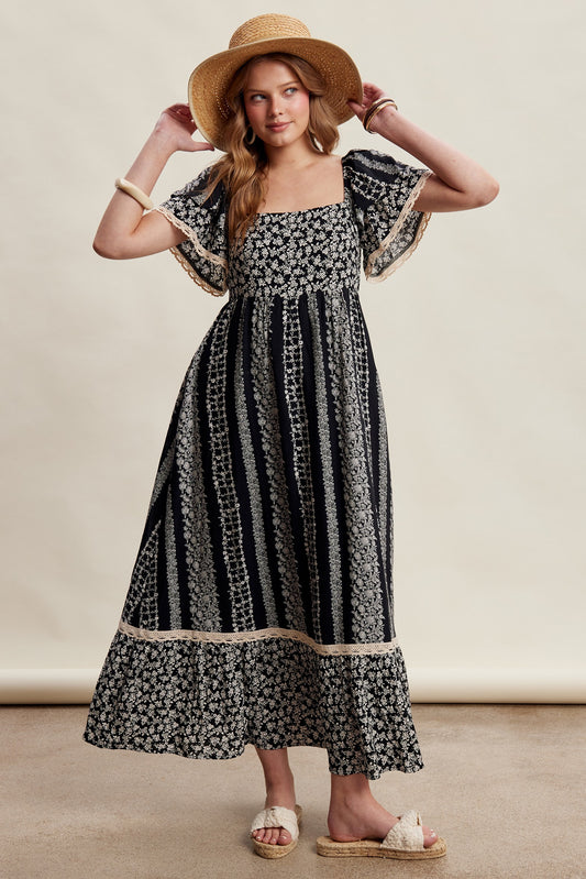 A black floral border print maxi dress featuring a square neckline, smocked bodice, tiered flutter sleeves with lace trim, and functional side pockets.