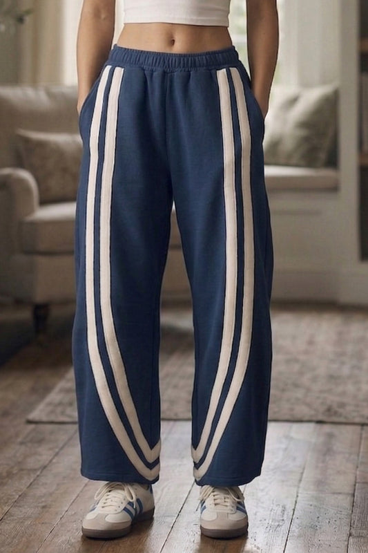 Navy blue barrel-leg track pants featuring ivory side stripe detailing, a high-rise elastic waistband, functional side pockets, and a relaxed curved-leg silhouette.