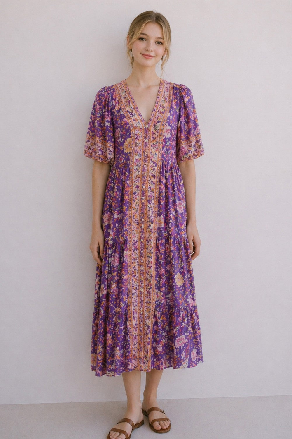 A short-sleeve tiered floral maxi dress in a lavender, coral, and gold mix featuring a deep V-neckline, button-down front closure, and decorative border print detailing.
