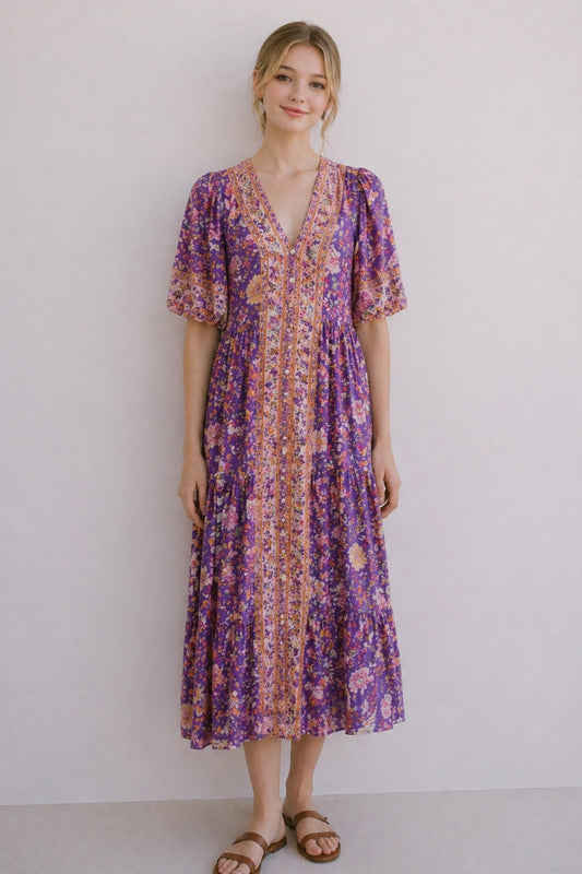 A short-sleeve tiered floral maxi dress in a lavender, coral, and gold mix featuring a deep V-neckline, button-down front closure, and decorative border print detailing.