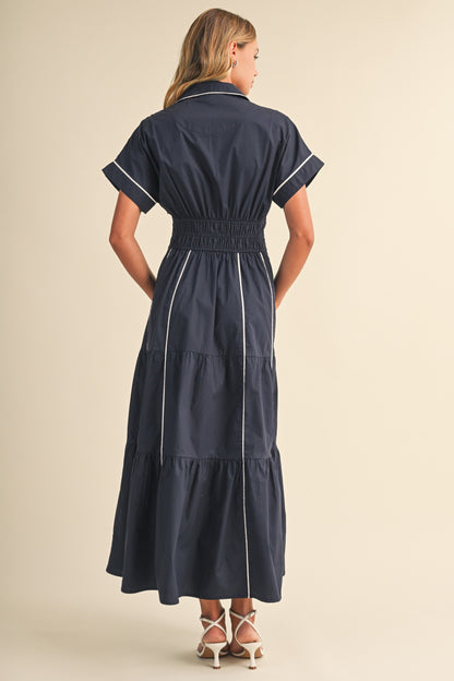 A short-sleeve navy blue shirt maxi dress featuring a classic collar, white vertical contrast piping, a thick smocked elastic waistband, and a tiered A-line skirt.