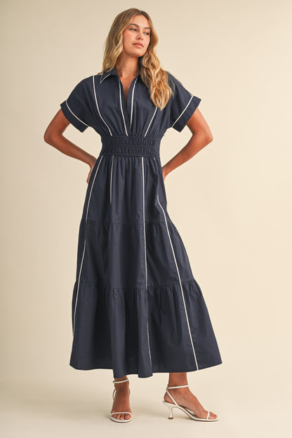 A short-sleeve navy blue shirt maxi dress featuring a classic collar, white vertical contrast piping, a thick smocked elastic waistband, and a tiered A-line skirt.