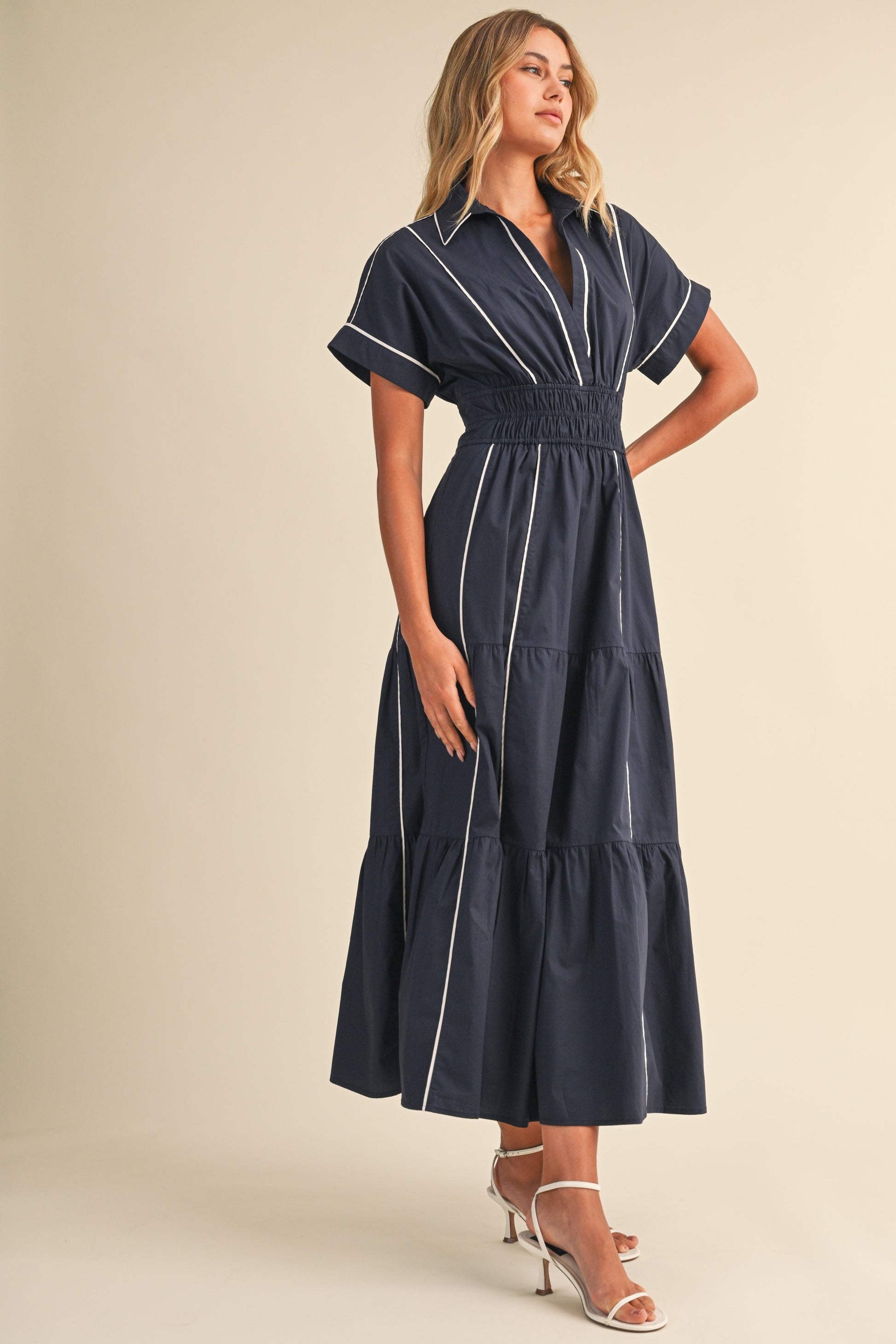 A short-sleeve navy blue shirt maxi dress featuring a classic collar, white vertical contrast piping, a thick smocked elastic waistband, and a tiered A-line skirt.