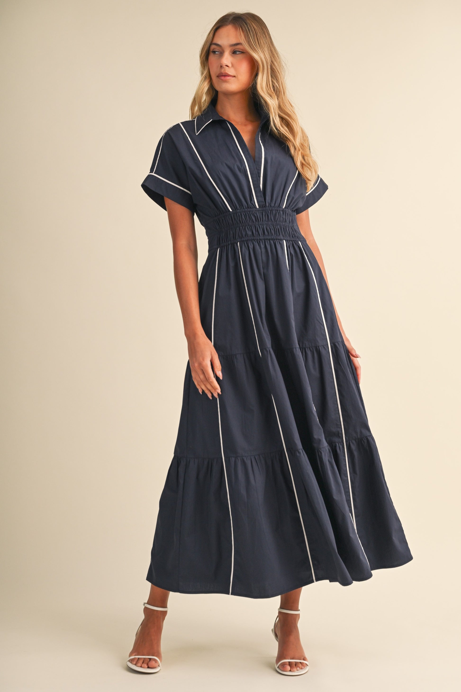 A short-sleeve navy blue shirt maxi dress featuring a classic collar, white vertical contrast piping, a thick smocked elastic waistband, and a tiered A-line skirt.