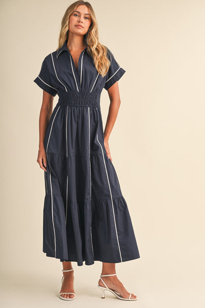 A short-sleeve navy blue shirt maxi dress featuring a classic collar, white vertical contrast piping, a thick smocked elastic waistband, and a tiered A-line skirt.
