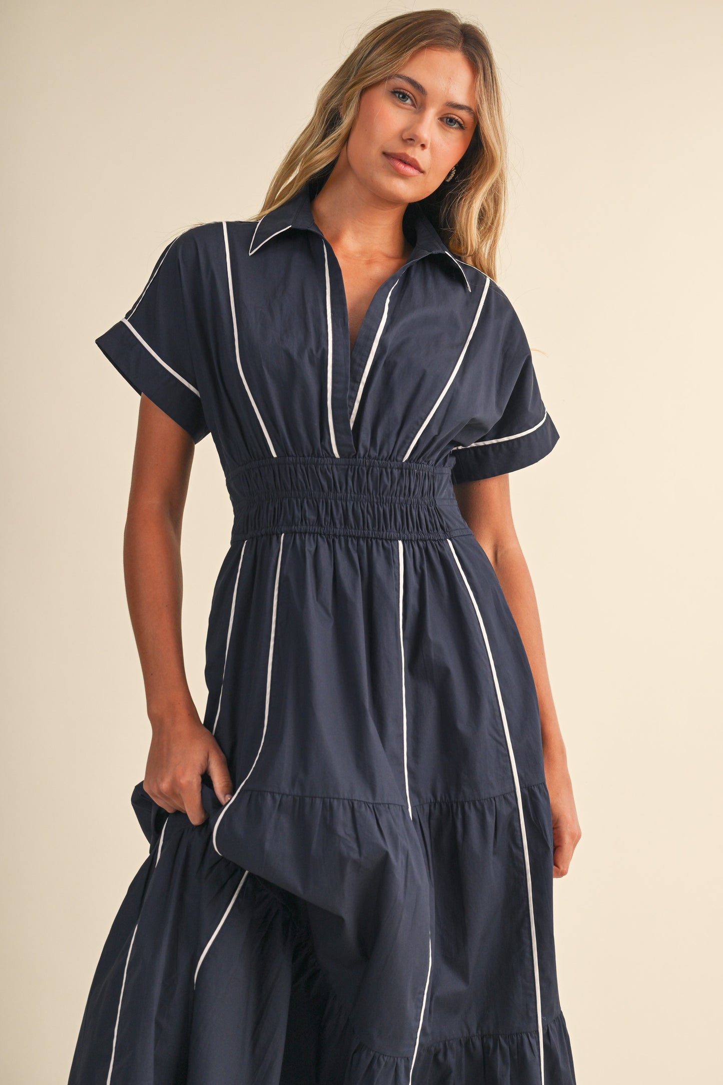 A short-sleeve navy blue shirt maxi dress featuring a classic collar, white vertical contrast piping, a thick smocked elastic waistband, and a tiered A-line skirt.