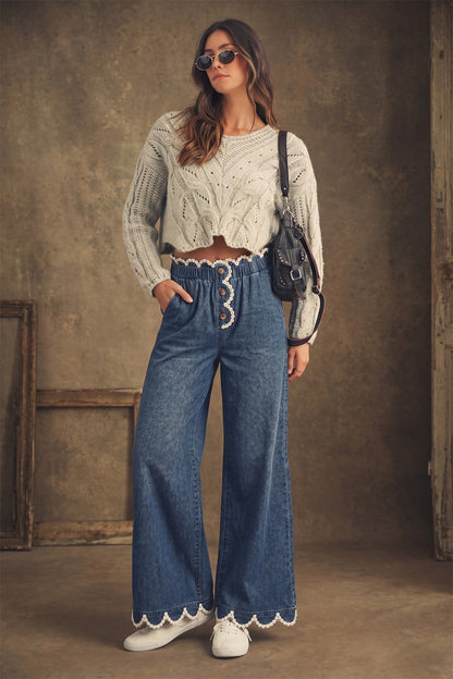 A pair of women's high-rise draped wide-leg denim pants in a deep indigo wash featuring a gathered elastic paperbag waistband, a button-front fly, functional side pockets, and a white daisy-edge scallop lace trim along the waist and bottom hems.