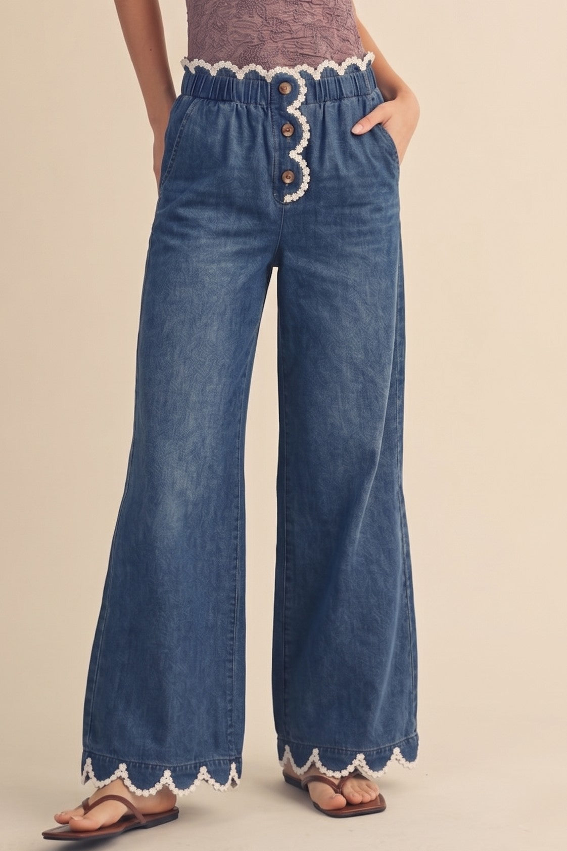 A pair of women's high-rise draped wide-leg denim pants in a deep indigo wash featuring a gathered elastic paperbag waistband, a button-front fly, functional side pockets, and a white daisy-edge scallop lace trim along the waist and bottom hems.