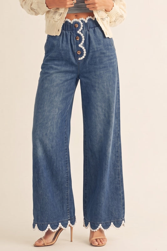 A pair of women's high-rise draped wide-leg denim pants in a deep indigo wash featuring a gathered elastic paperbag waistband, a button-front fly, functional side pockets, and a white daisy-edge scallop lace trim along the waist and bottom hems.