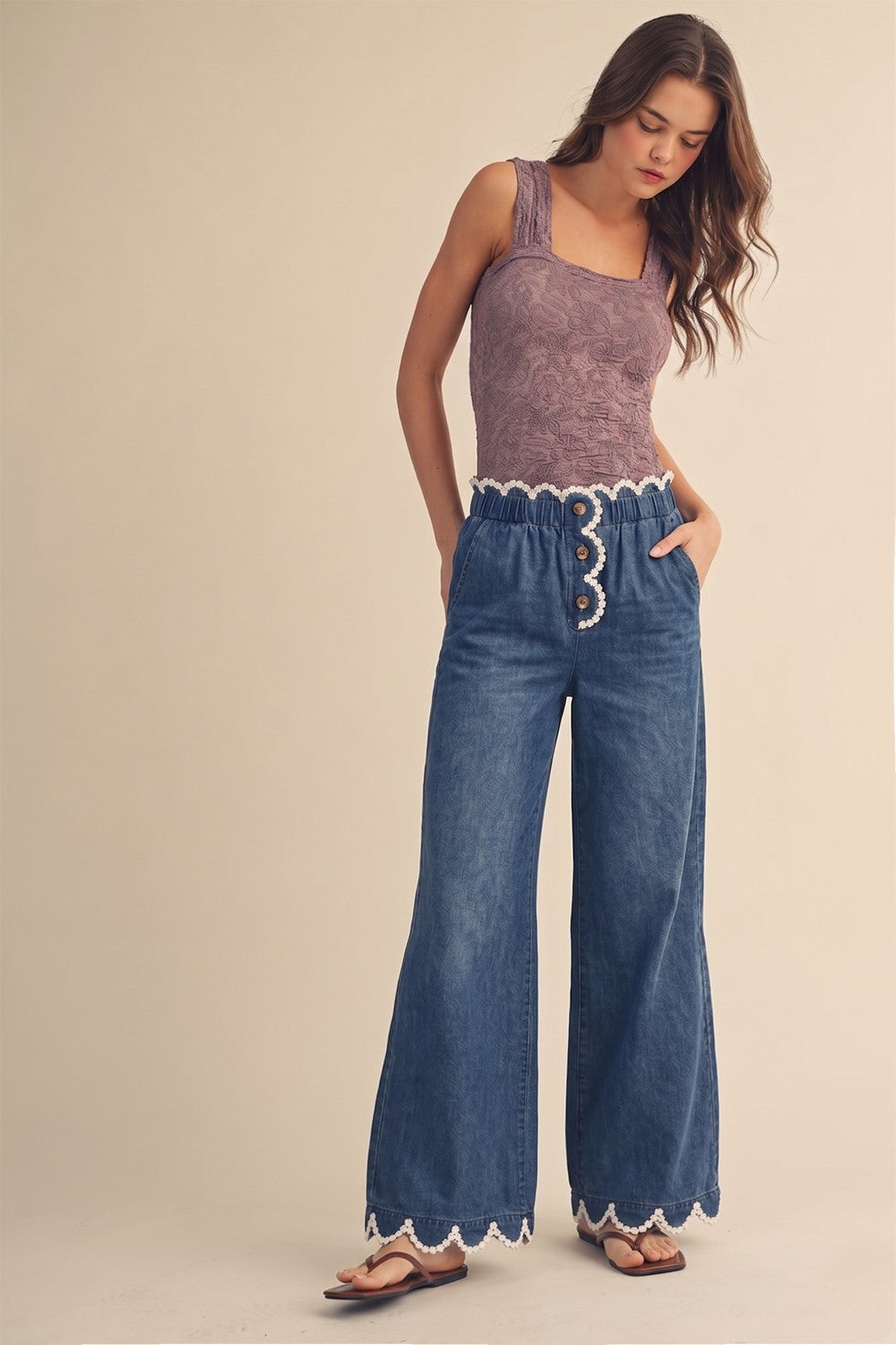 A pair of women's high-rise draped wide-leg denim pants in a deep indigo wash featuring a gathered elastic paperbag waistband, a button-front fly, functional side pockets, and a white daisy-edge scallop lace trim along the waist and bottom hems.