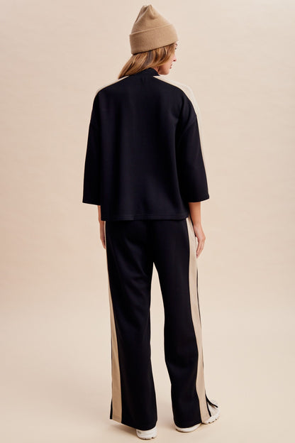 Women's two-piece black lounge set featuring a half-sleeve mock-neck top, neutral side-stripe detailing, and matching wide-leg pants with ankle hem slits.