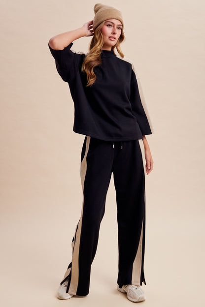 Women's two-piece black lounge set featuring a half-sleeve mock-neck top, neutral side-stripe detailing, and matching wide-leg pants with ankle hem slits.