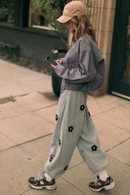 A women's heather gray wide-leg fleece sweatpants featuring an elastic drawstring waistband, relaxed barrel leg, and black chenille flower patches across the front and back.