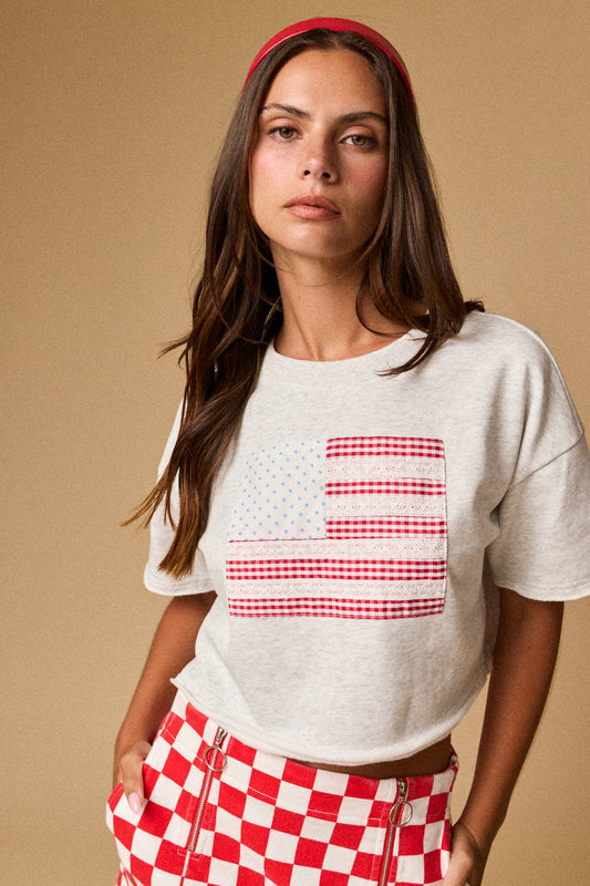 A women's light gray short-sleeve graphic t-shirt featuring a relaxed fit, a raw cut cropped hemline, and a textured patchwork American flag design made from blue star print, red gingham, and white eyelet lace fabrics.