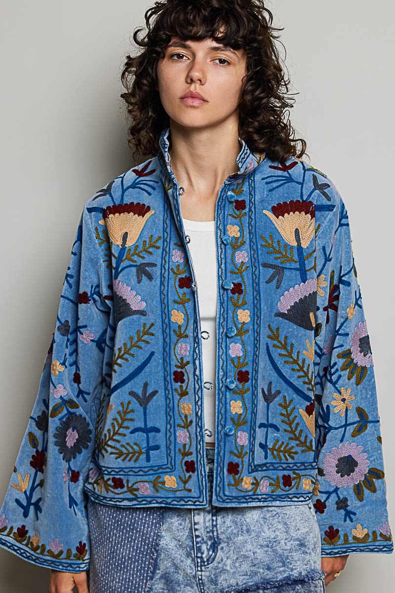 A women's dusty denim blue long-sleeve velvet jacket with allover folk floral embroidery in rust, yellow, and lavender, styled with a mandarin collar, wide cuffs, and front loop buttons.