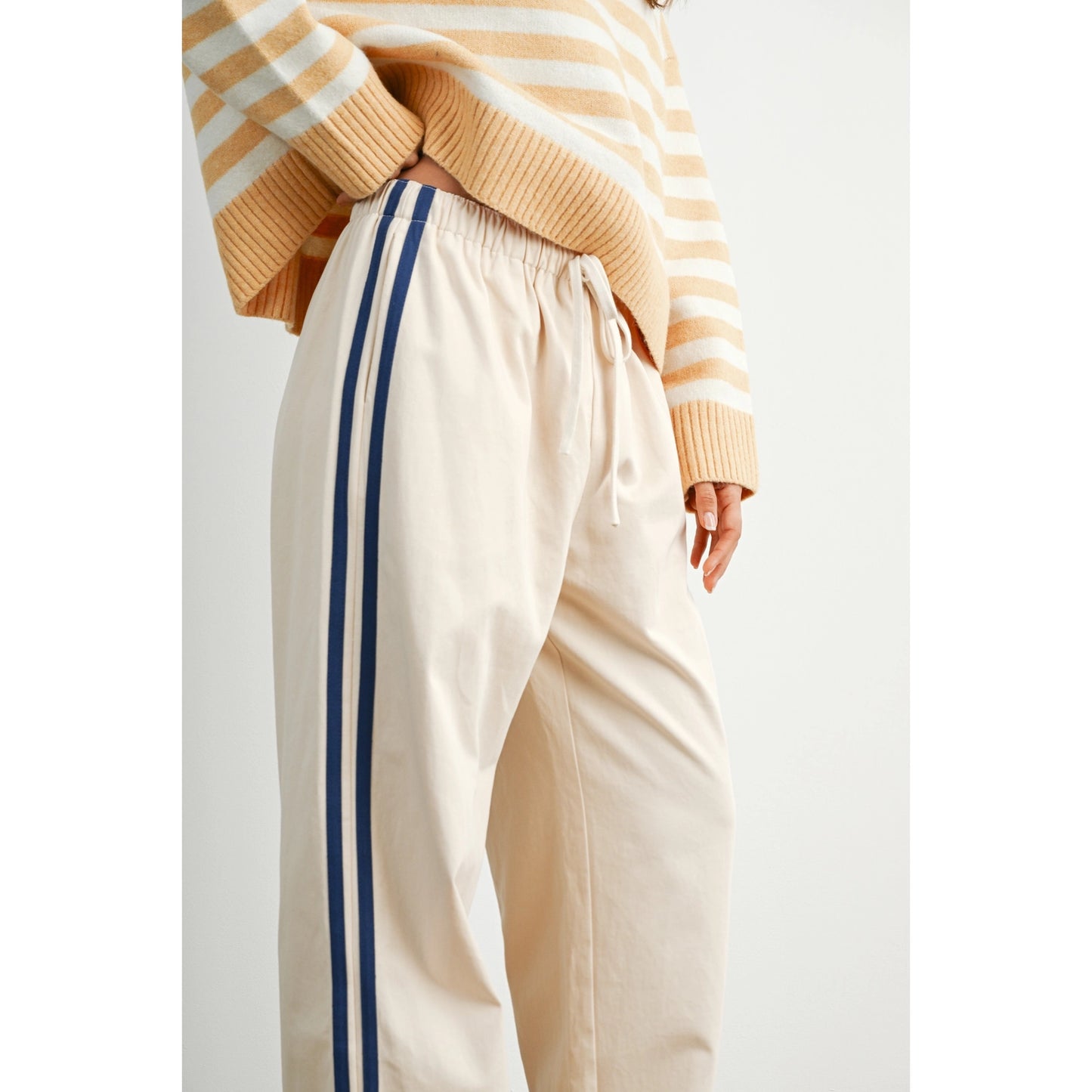 A pair of women's wide-leg relaxed cream track pants featuring double navy blue side stripes, an elastic drawstring waistband, and a mid-rise fit.