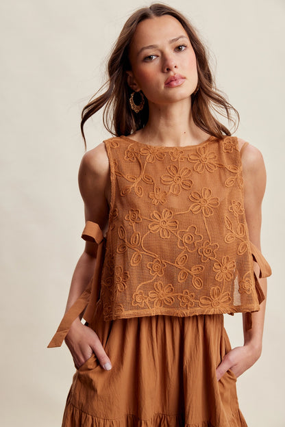 A women's brown sleeveless tiered maxi dress featuring a tonal embroidered floral lace vest overlay with adjustable side tie details.