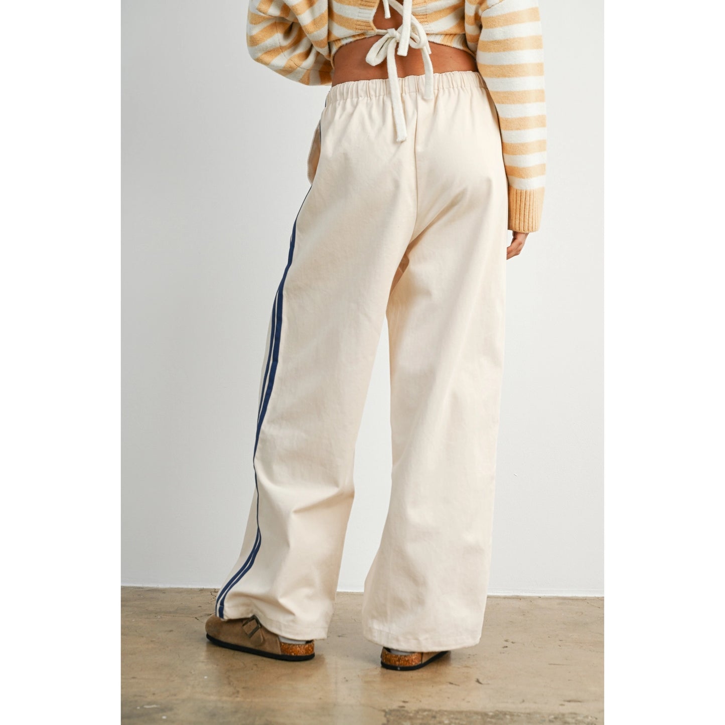 A pair of women's wide-leg relaxed cream track pants featuring double navy blue side stripes, an elastic drawstring waistband, and a mid-rise fit.