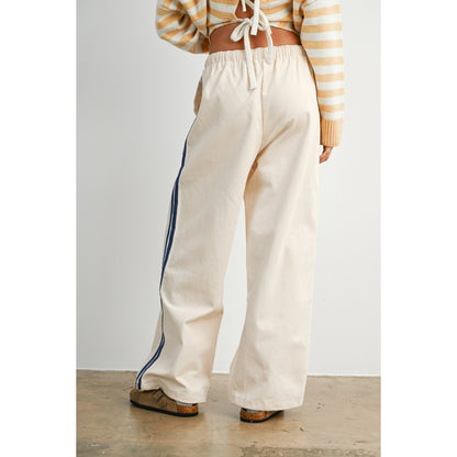 A pair of women's wide-leg relaxed cream track pants featuring double navy blue side stripes, an elastic drawstring waistband, and a mid-rise fit.