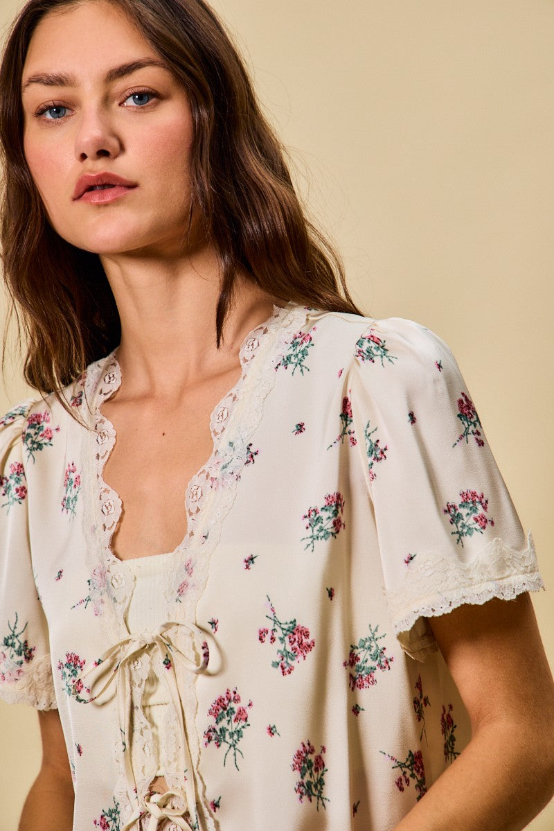 A short-sleeve ivory satin floral print blouse featuring a functional double tie-front closure, puff sleeves, and scalloped lace trim along the neckline and hem.