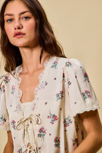 A short-sleeve ivory satin floral print blouse featuring a functional double tie-front closure, puff sleeves, and scalloped lace trim along the neckline and hem.