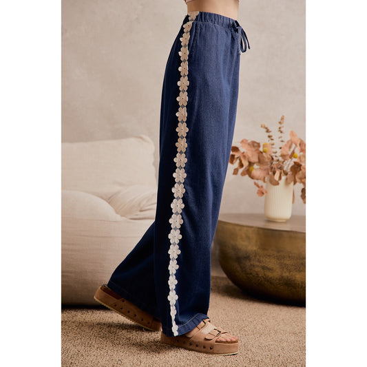 A women's medium wash wide-leg denim pants featuring a gathered elastic drawstring waistband and white crochet daisy floral lace side seam trim.