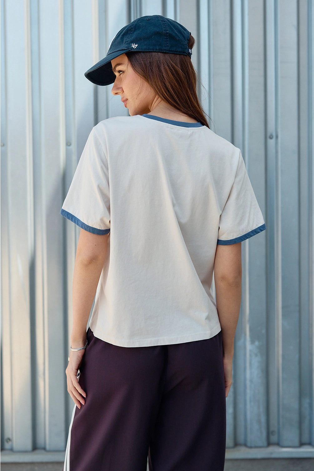 A women's light sand crewneck ringer t-shirt featuring deep blue contrast trim along the neckline and sleeves, short sleeves, and cursive blue embroidered script across the chest that says, Merci. 