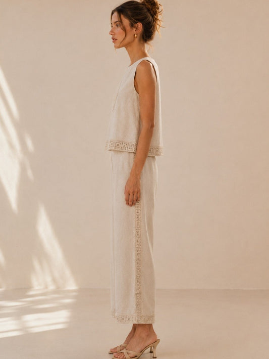 A women's two-piece linen-blend clothing set in an oat color featuring a sleeveless top with a chest diamond crochet applique and matching high-waisted wide-leg pants with lace trim panels along the sides and hems.