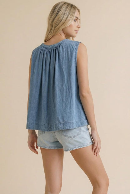 A sleeveless washed blue denim top featuring a functional button-down front, a rounded neckline with shirred detailing, and a relaxed swing silhouette.