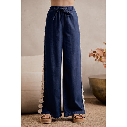 A women's medium wash wide-leg denim pants featuring a gathered elastic drawstring waistband and white crochet daisy floral lace side seam trim.