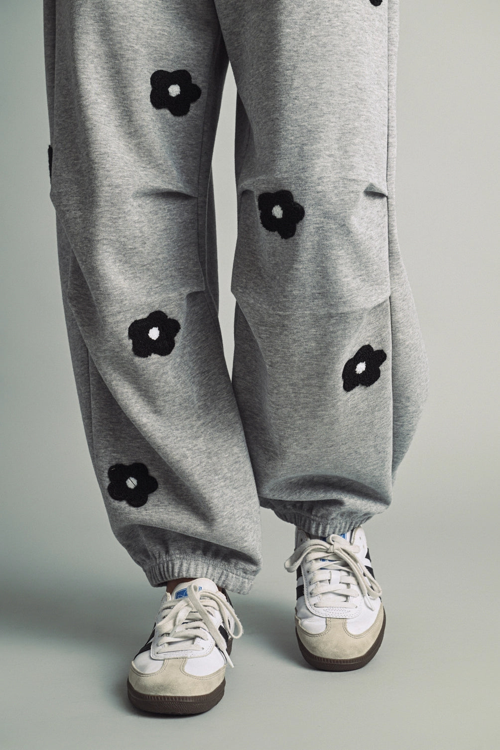 A women's heather gray wide-leg fleece sweatpants featuring an elastic drawstring waistband, relaxed barrel leg, and black chenille flower patches across the front and back.