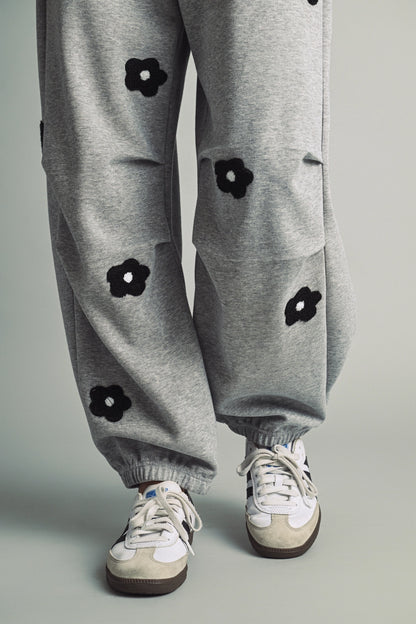 A women's heather gray wide-leg fleece sweatpants featuring an elastic drawstring waistband, relaxed barrel leg, and black chenille flower patches across the front and back.