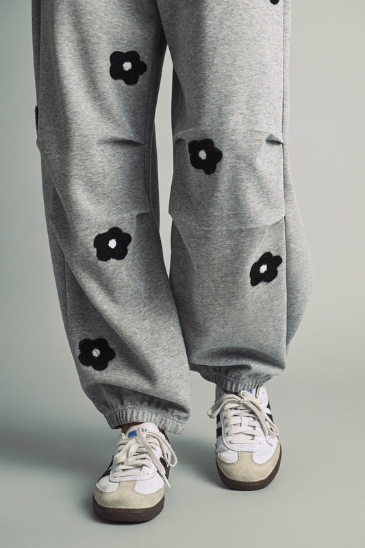 A women's heather gray wide-leg fleece sweatpants featuring an elastic drawstring waistband, relaxed barrel leg, and black chenille flower patches across the front and back.