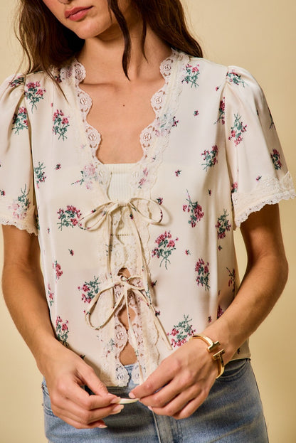 A short-sleeve ivory satin floral print blouse featuring a functional double tie-front closure, puff sleeves, and scalloped lace trim along the neckline and hem.