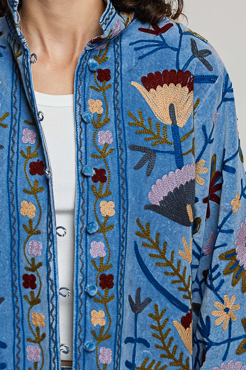 A women's dusty denim blue long-sleeve velvet jacket with allover folk floral embroidery in rust, yellow, and lavender, styled with a mandarin collar, wide cuffs, and front loop buttons.