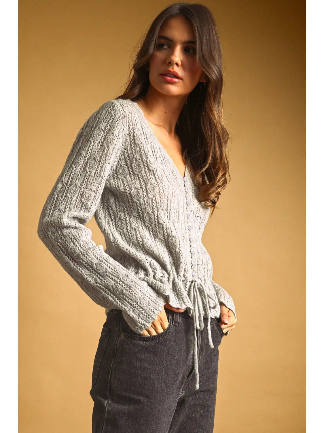 A women's light gray long-sleeve textured lace knit cardigan sweater featuring a button-down front, scalloped V-neckline, and an adjustable drawstring tie waistline.