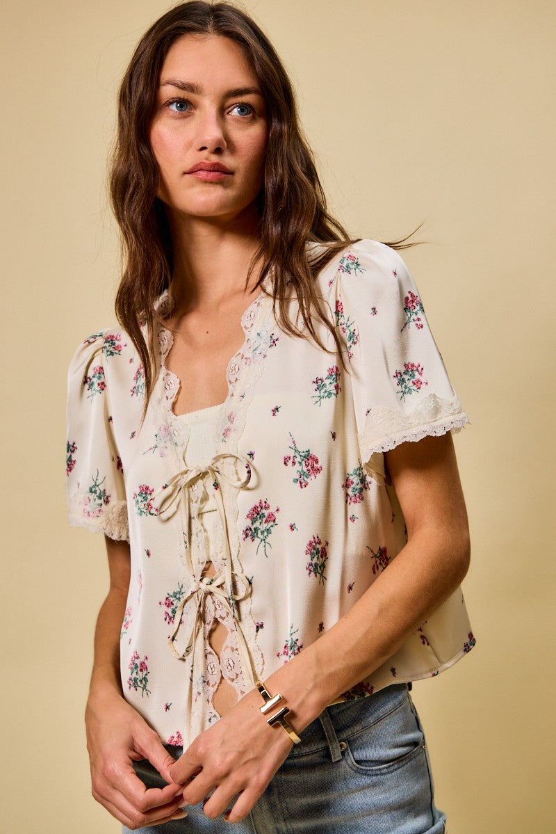 A short-sleeve ivory satin floral print blouse featuring a functional double tie-front closure, puff sleeves, and scalloped lace trim along the neckline and hem.