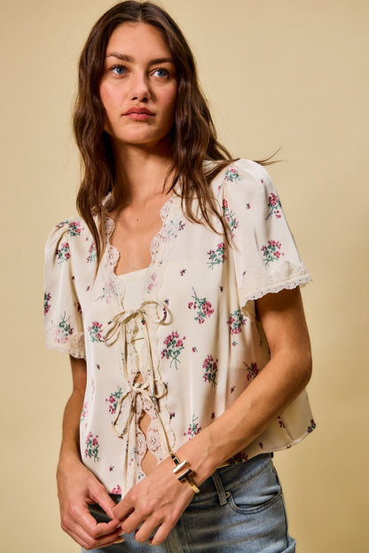 A short-sleeve ivory satin floral print blouse featuring a functional double tie-front closure, puff sleeves, and scalloped lace trim along the neckline and hem.