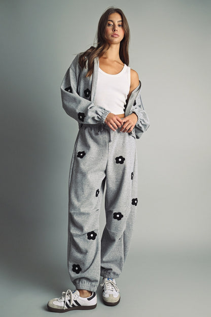 A women's heather gray wide-leg fleece sweatpants featuring an elastic drawstring waistband, relaxed barrel leg, and black chenille flower patches across the front and back.
