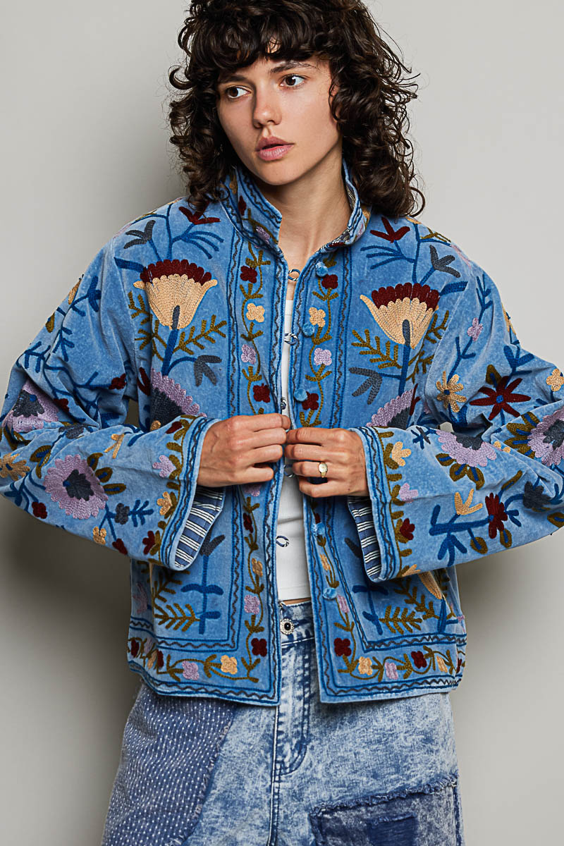 A women's dusty denim blue long-sleeve velvet jacket with allover folk floral embroidery in rust, yellow, and lavender, styled with a mandarin collar, wide cuffs, and front loop buttons.