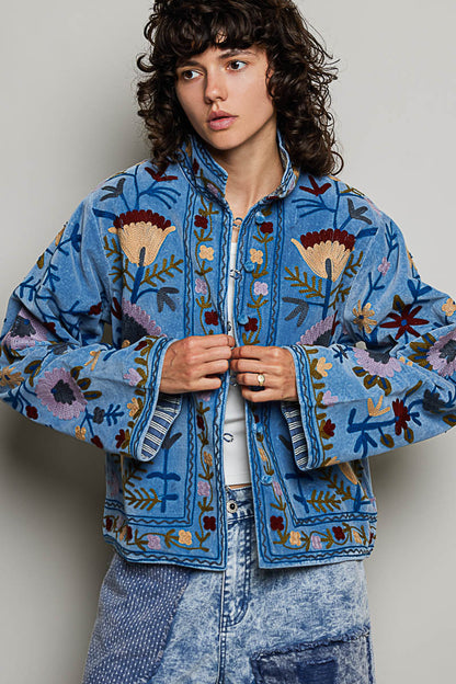 A women's dusty denim blue long-sleeve velvet jacket with allover folk floral embroidery in rust, yellow, and lavender, styled with a mandarin collar, wide cuffs, and front loop buttons.