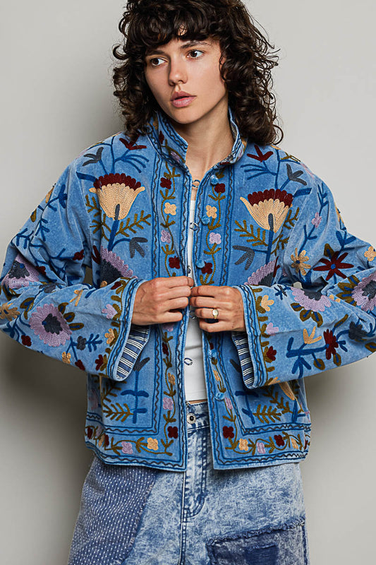 A women's dusty denim blue long-sleeve velvet jacket with allover folk floral embroidery in rust, yellow, and lavender, styled with a mandarin collar, wide cuffs, and front loop buttons.