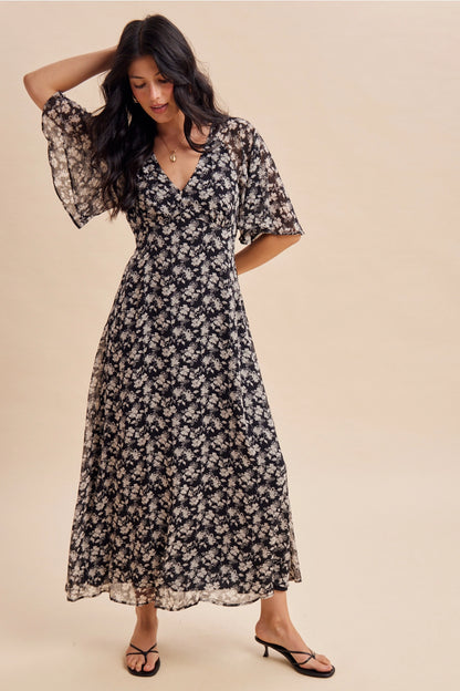A women's v-neck maxi dress in a black and cream floral print featuring short flutter sleeves and an empire waist silhouette.