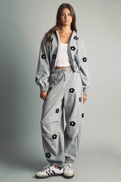 A women's heather gray full-zip hooded fleece jacket featuring dropped shoulders, ribbed hem cuffs, and black chenille flower patches on the front and sleeves.